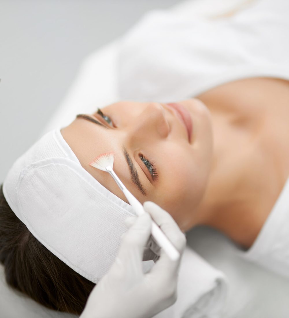 Side view of beautician holding brush and doing cleaning face for young beautiful woman. Concept of procedure for rejuvenation skin or improvements in professional beauty salon.