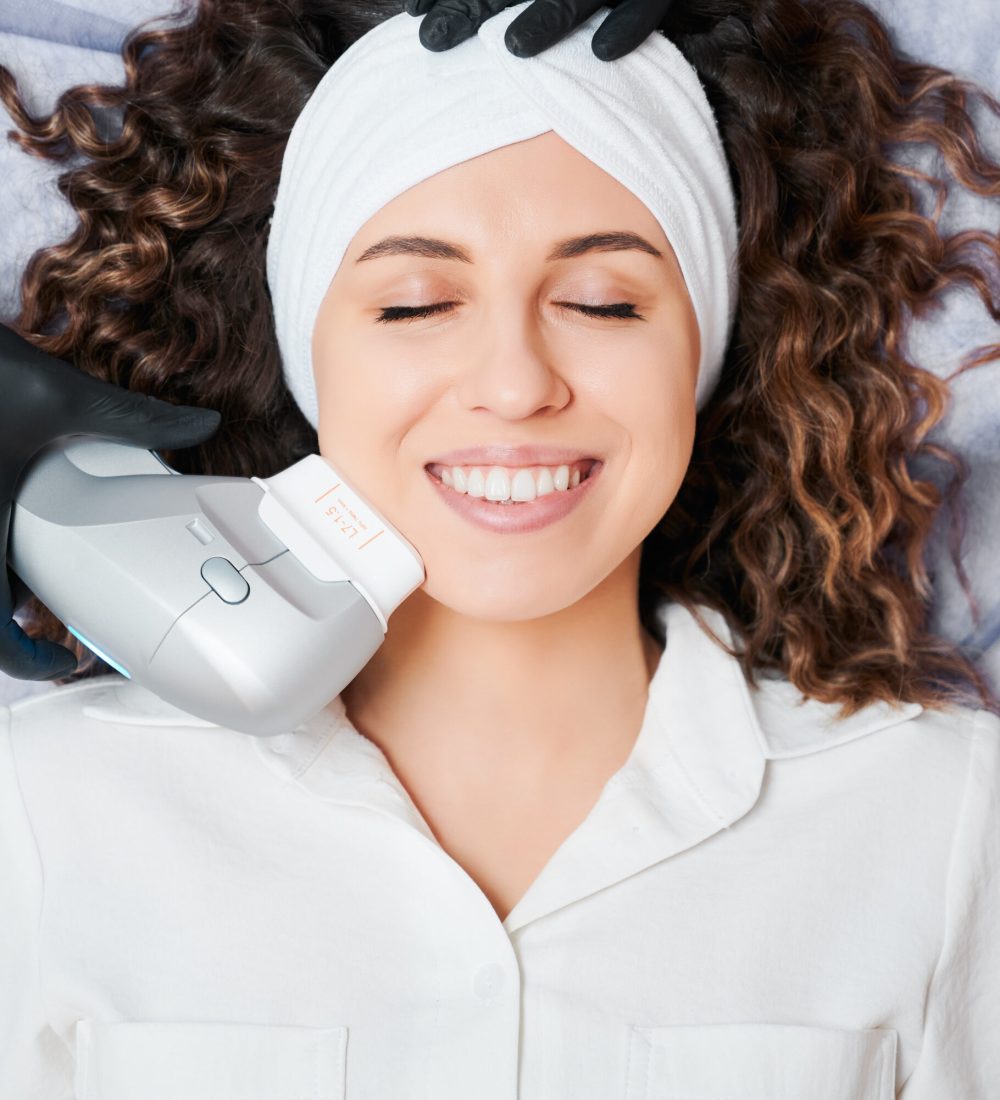 Top view of beautiful young woman with closed eyes wearing white headband while receiving facial treatment at wellness center. Cheerful female patient having face lifting procedure and smiling.