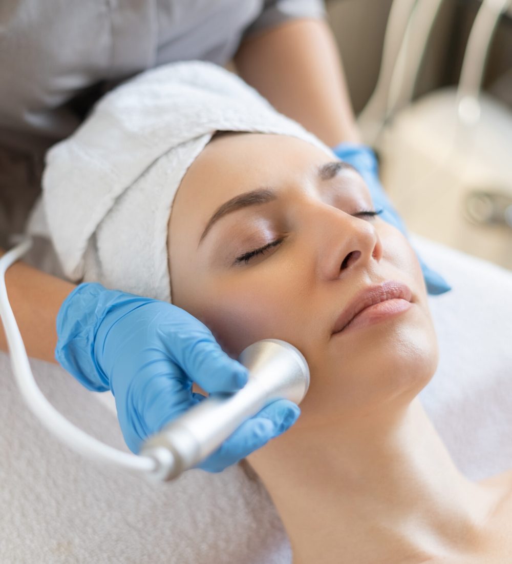 Young woman lying on cosmetologist's table during rejuvenation procedure. Cosmetologist take care about neck and face skin youthfull and wellness. Hardware face cleaning procedure